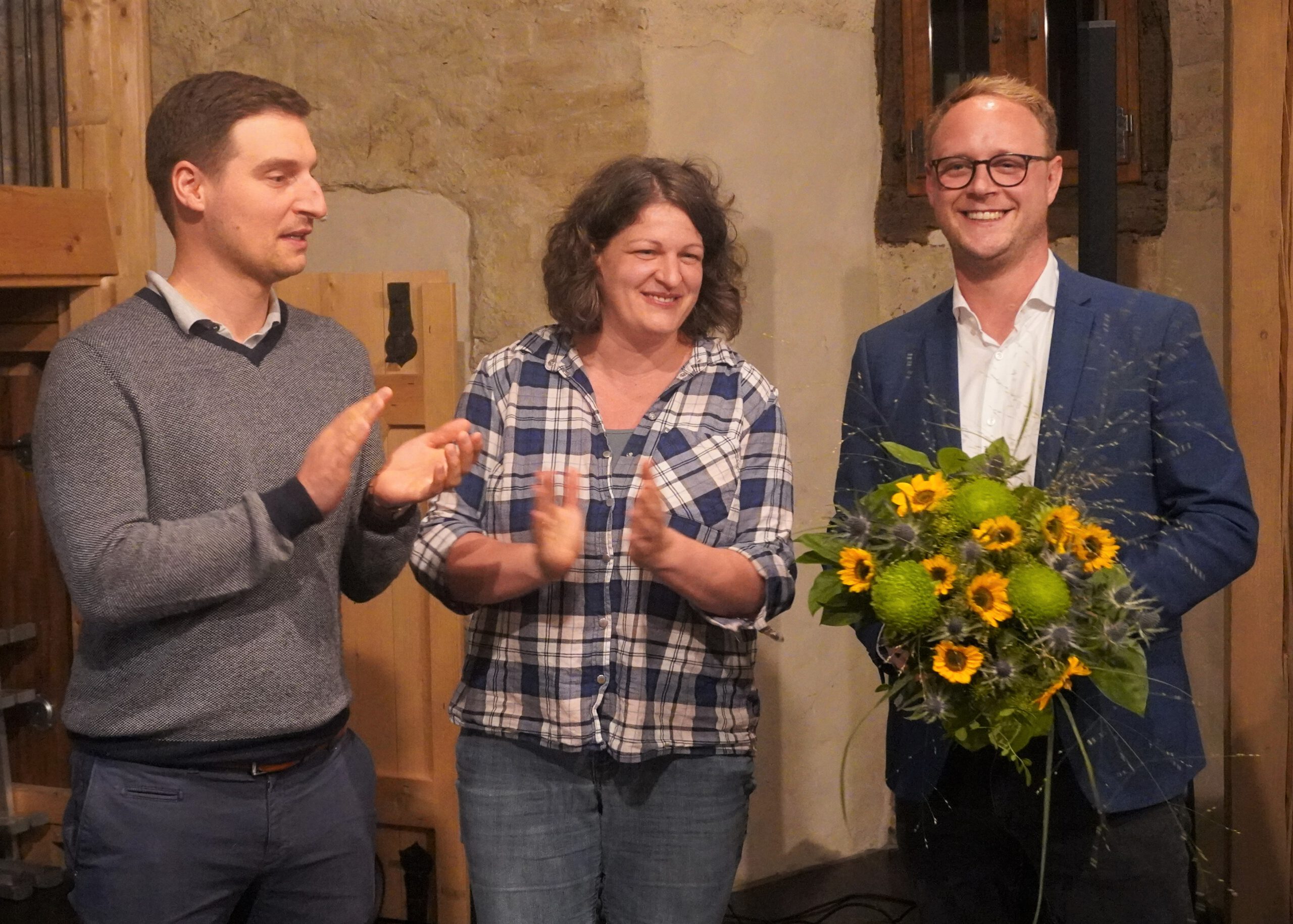 Nils Lundt, Anna Miehe und Marcel Emmerich sind zu sehen. Marcel Emmerich hält einen Blumenstrauß, Anna Miehe und Nils Lundt applaudieren.